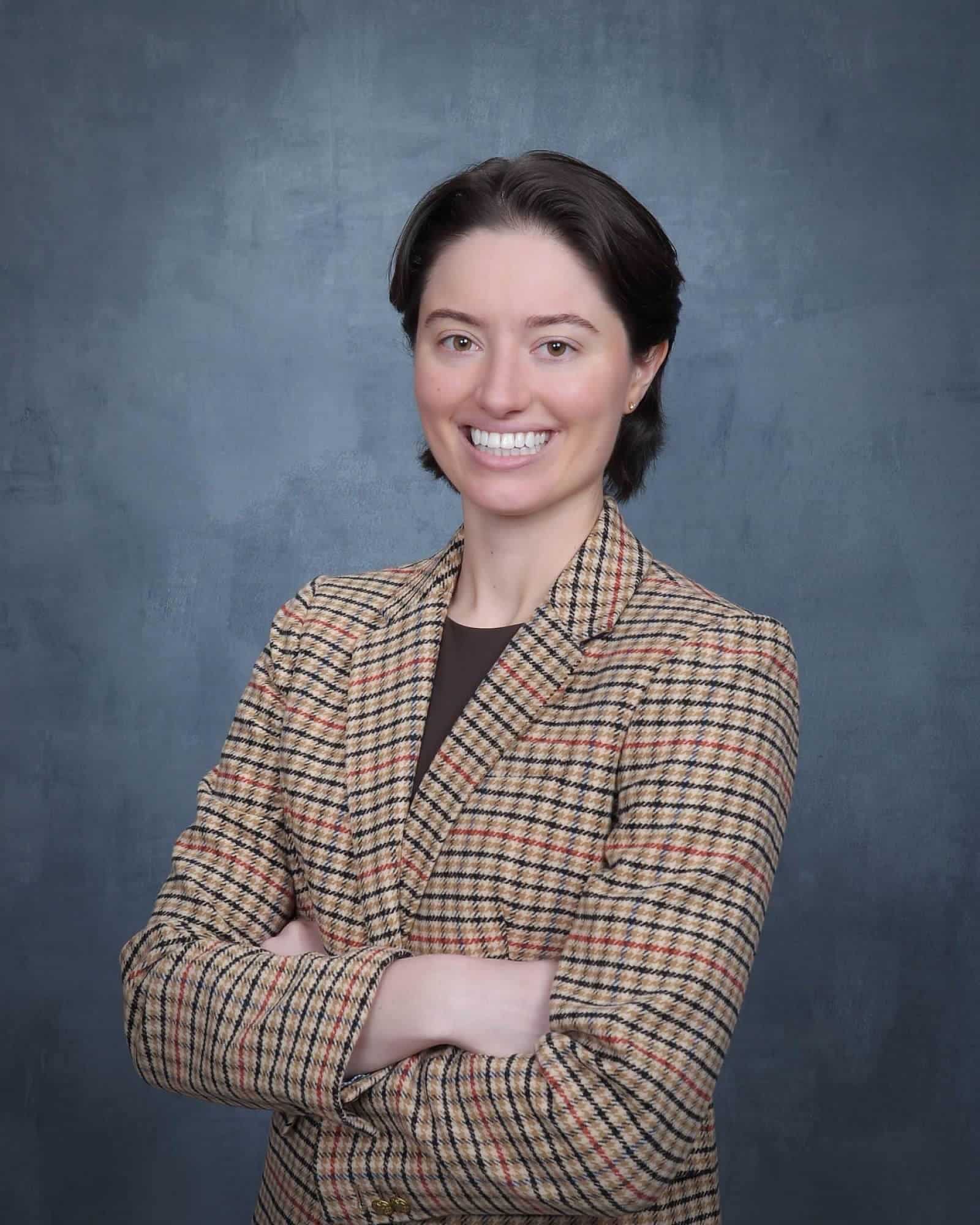 Eleanor Bullock, MSN, APRN, FNP-BC, smiling with arms folded, in a check blazer against a grey studio backdrop